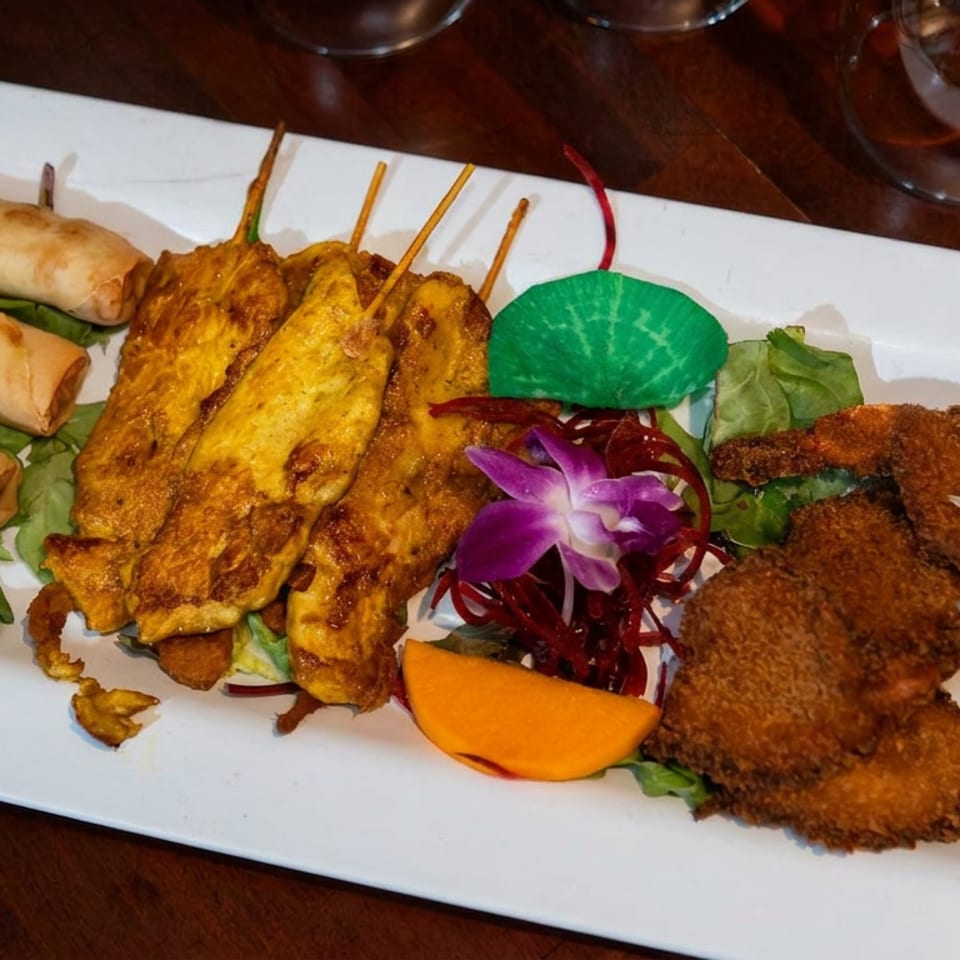Assorted Asian appetizers on a white platter, including spring rolls, satay skewers, fried seafood, greens, and decorative flowers.