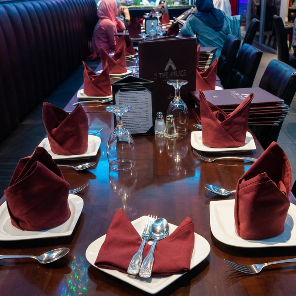 Restaurant dining table set with burgundy folded napkins, white plates, silverware, upside-down glasses, and menus beside a booth.