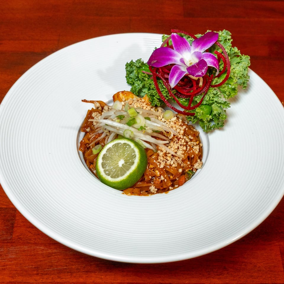 Thai-style noodles topped with bean sprouts, peanuts, lime, scallions, kale, beet strips, and an orchid garnish on a white plate.