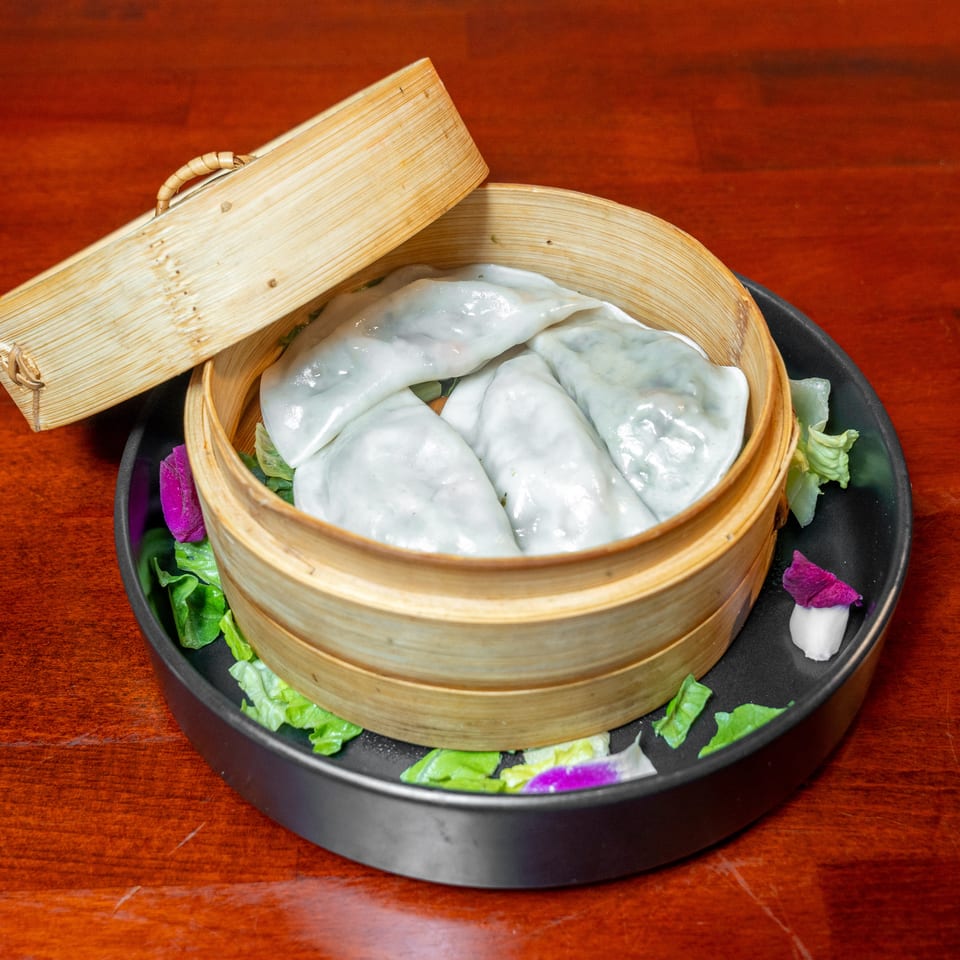 Steamed dumplings in an open bamboo basket, garnished with lettuce and purple edible flowers on a black plate.