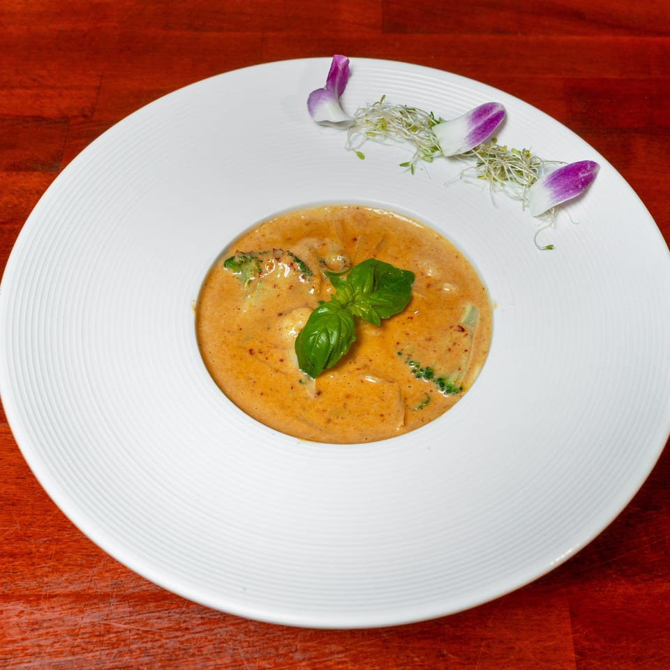 Creamy orange curry garnished with fresh basil, broccoli, sprouts, and purple flower petals on a white plate.
