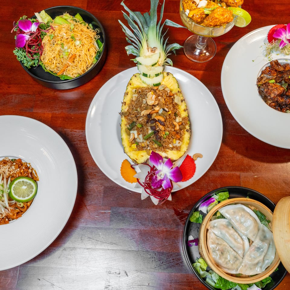 Overhead spread of Asian dishes including pineapple fried rice, noodles, dumplings, glazed meat, and garnished plates on a wooden table.
