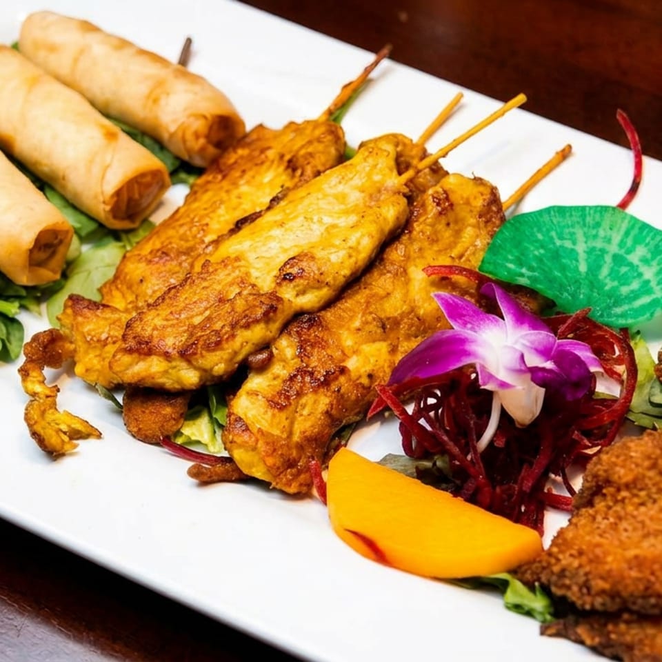 Assorted Asian appetizers on a white platter, including spring rolls, grilled skewers, fried bites, greens, mango, and orchid garnish.