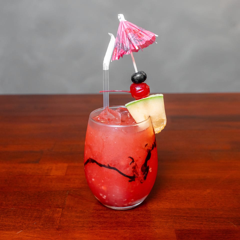 Red iced cocktail garnished with a melon wedge, cherries, straw, and pink paper umbrella on a wooden table.