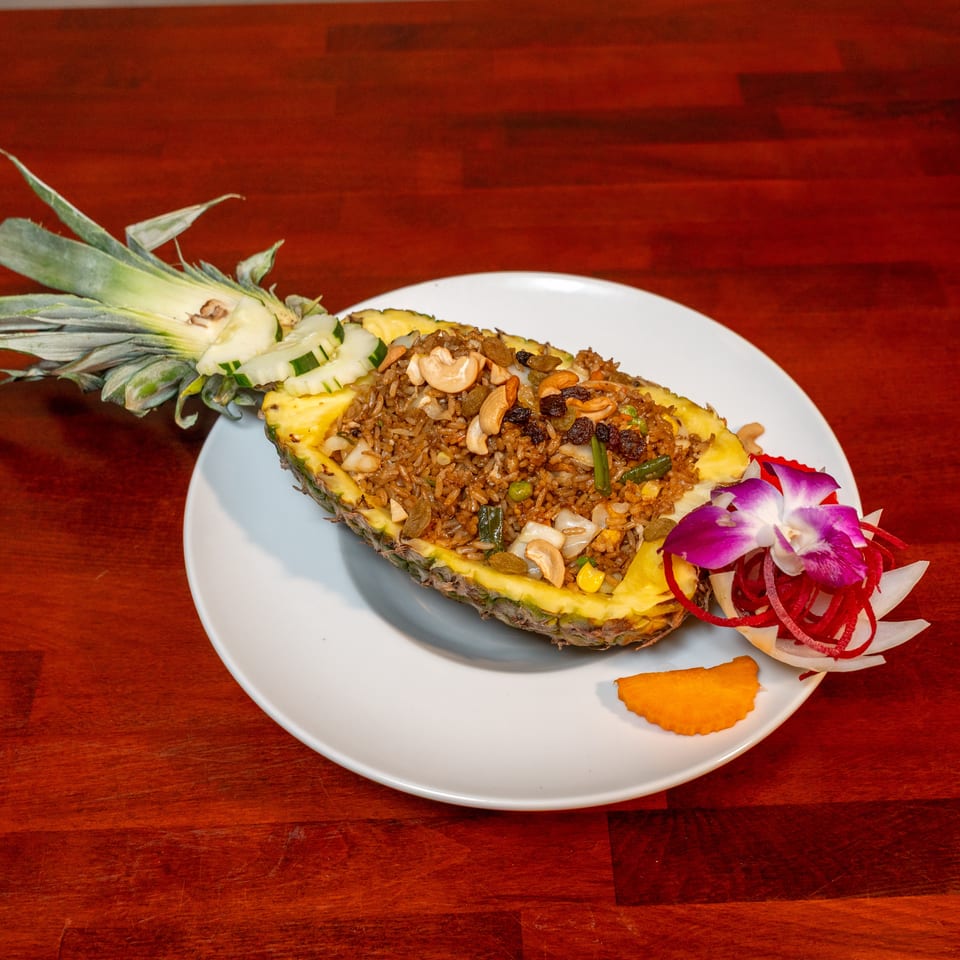 Fried rice with cashews, raisins, and vegetables served in a pineapple half on a white plate, garnished with flowers.