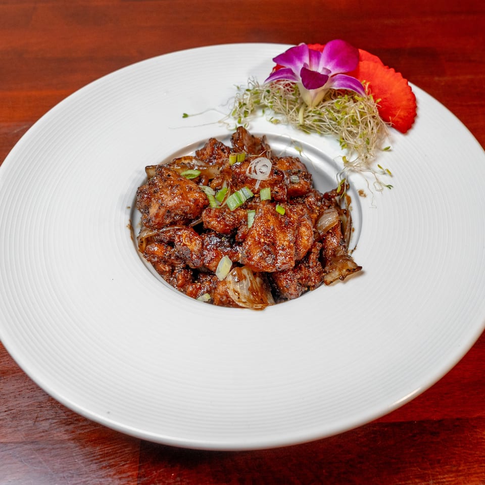 Glazed grilled meat topped with scallions, served on a white plate with sprouts and edible flowers.