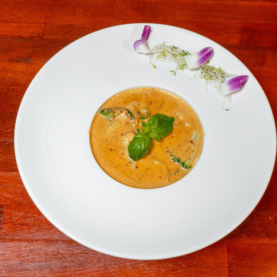 Creamy orange curry garnished with fresh basil, sprouts, and purple flower petals on a white plate atop a wooden table.