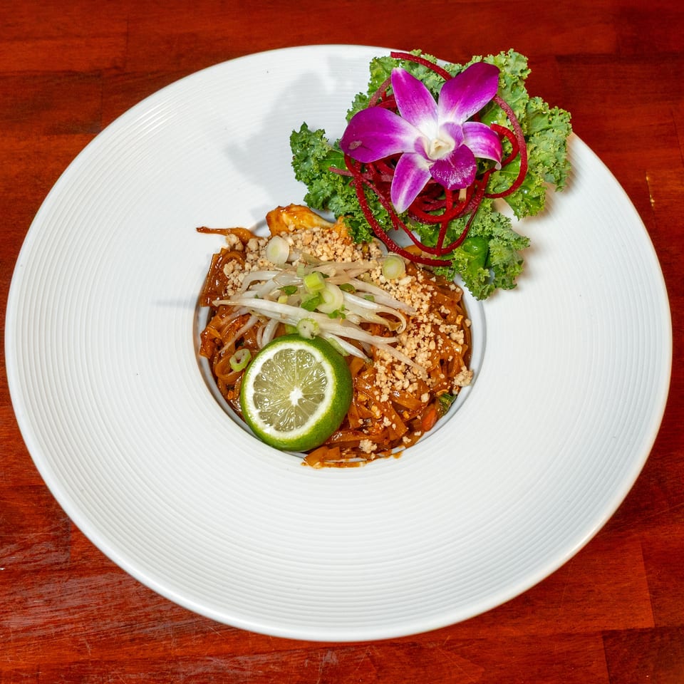 Thai-style noodles topped with bean sprouts, peanuts, lime, scallions, kale, and an orchid garnish on a white plate.