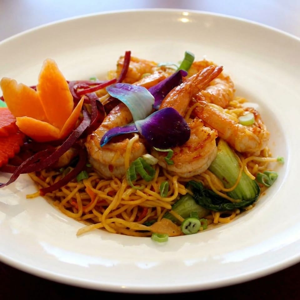 Plate of stir-fried noodles topped with shrimp, vegetables, purple cabbage, and colorful carved vegetable garnishes.