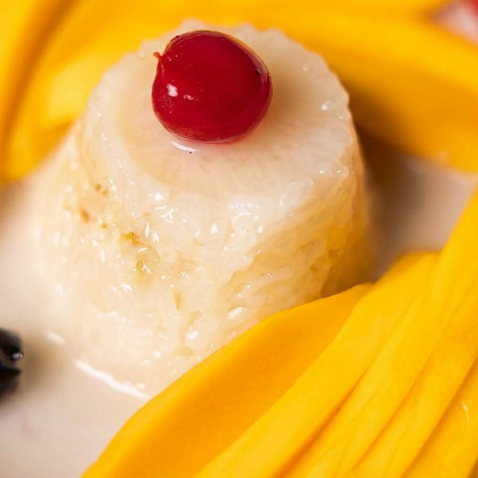 Glazed coconut dessert topped with a cherry, surrounded by sliced mango, banana, strawberries, and blueberries.