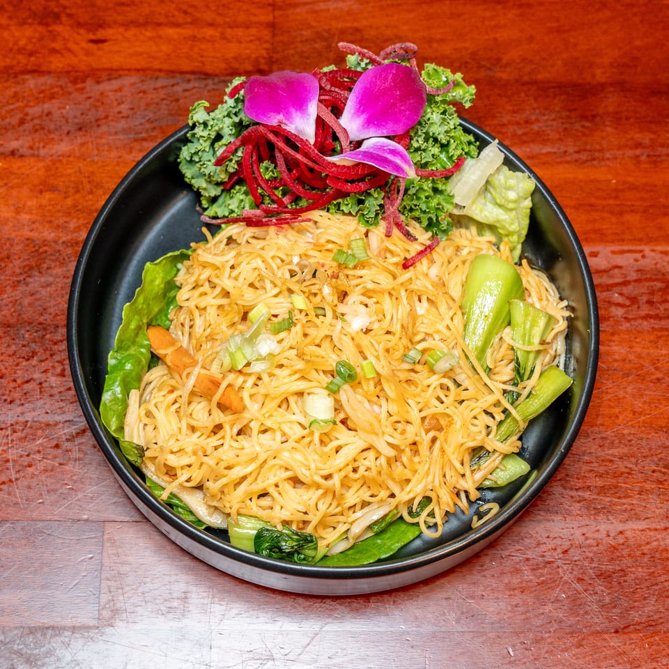 Stir-fried noodles with mixed vegetables, leafy greens, shredded beetroot, and purple radish petals in a black bowl on a wooden table.