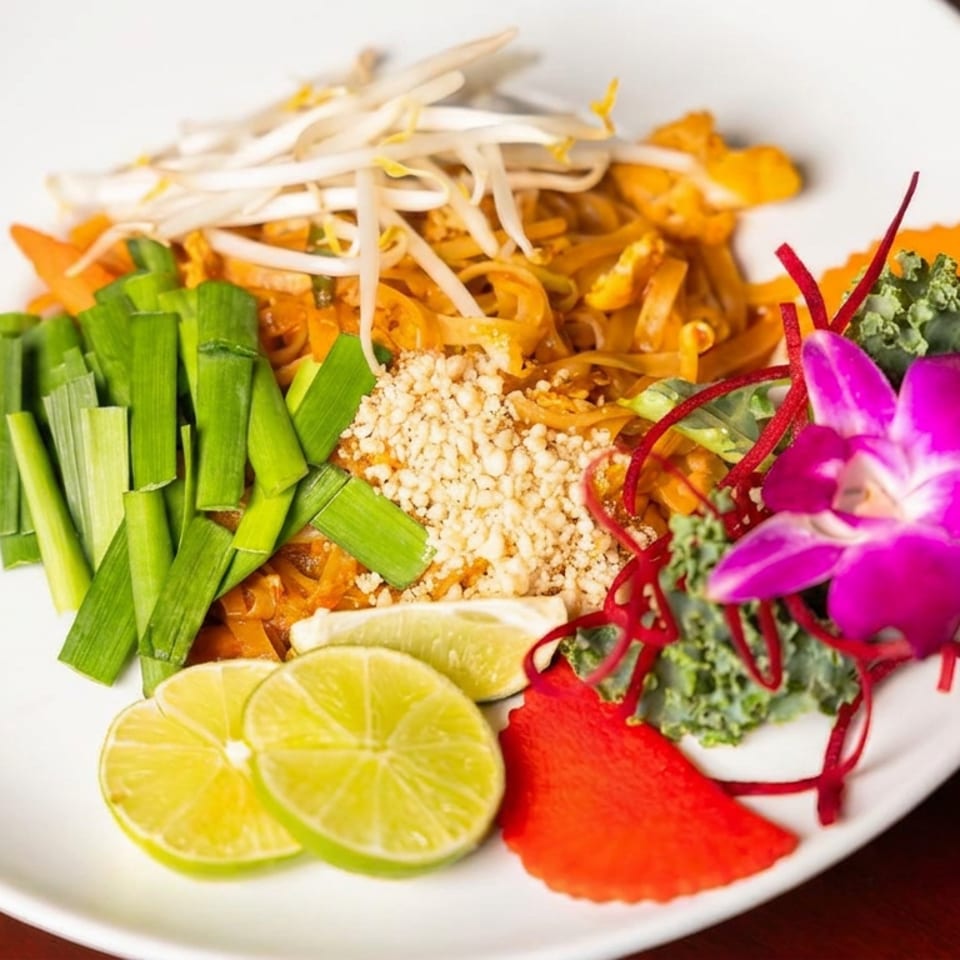 Plate of Thai-style noodles topped with bean sprouts, green onions, crushed peanuts, lime slices, vegetables, and a purple orchid garnish.