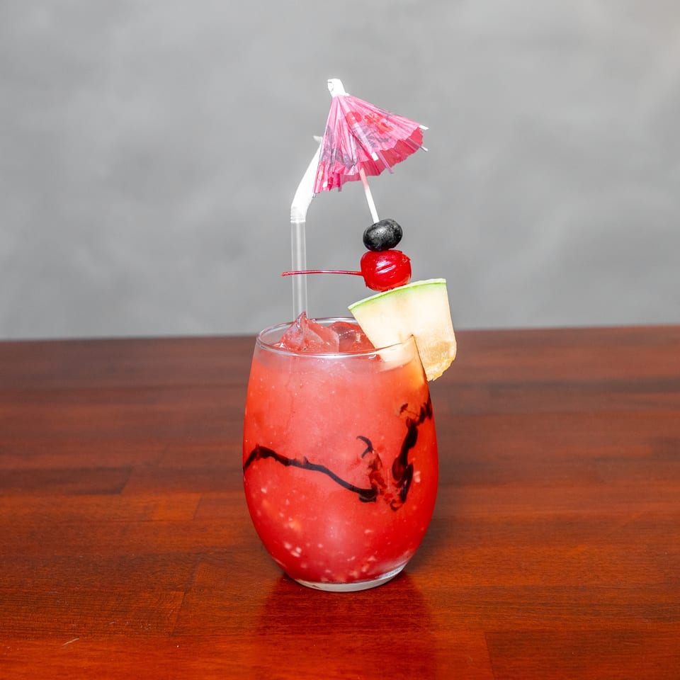 Red iced cocktail garnished with melon, cherries, and a pink paper umbrella on a wooden restaurant table.