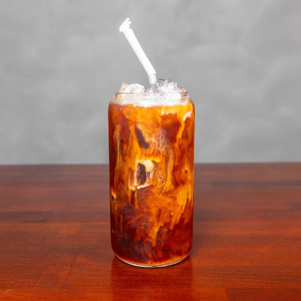 Iced amber beverage with cream swirling through it, served in a glass with a white straw on a wooden table.