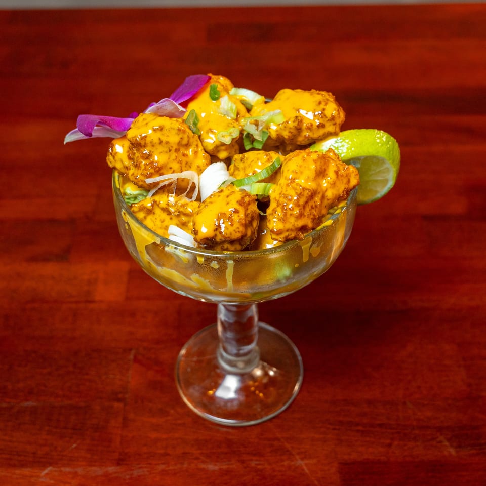 Crispy golden bites coated in creamy orange sauce, garnished with scallions, edible flowers, and lime in a cocktail glass.