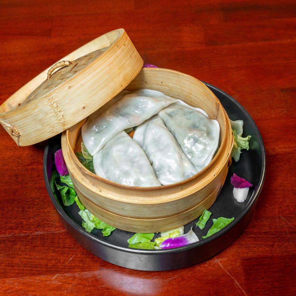 Steamed dumplings arranged in a bamboo basket with lettuce and purple garnishes on a black plate.